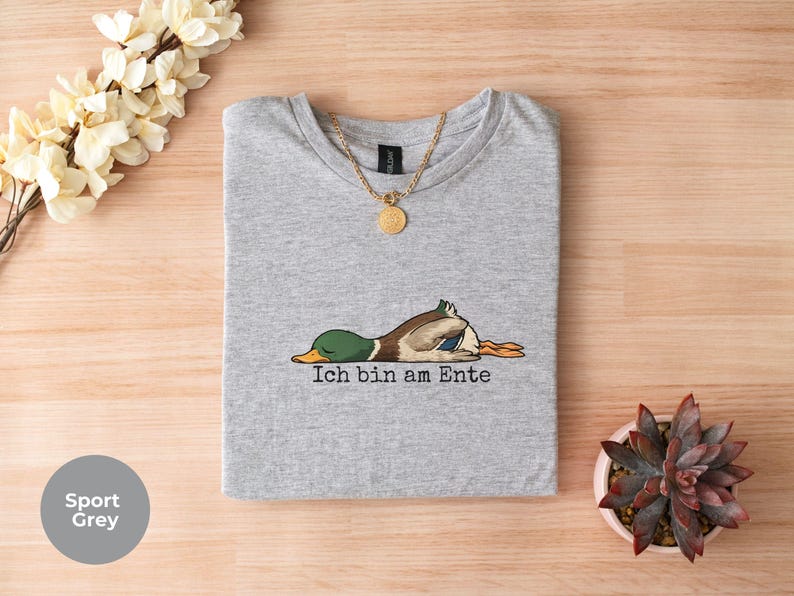 Pode incluir: Uma t-shirt cinzento desportivo com um gr&aacute;fico de um pato-real deitado de barriga para baixo e o texto Ich bin am Ente impresso a preto por baixo do pato. Um colar de corrente dourada repousa sobre o decote da t-shirt.