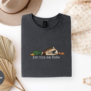 Pode incluir: Uma t-shirt cinzenta escura mesclada com um gr&aacute;fico de um pato-real deitado de barriga para baixo e o texto "Ich bin am Ente" impresso em branco por baixo da ilustra&ccedil;&atilde;o. A pe&ccedil;a tem gola redonda e mangas curtas.