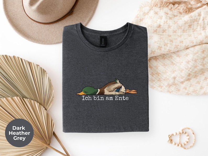 Pode incluir: Uma t-shirt cinzenta escura mesclada com um gr&aacute;fico de um pato-real deitado de barriga para baixo e o texto "Ich bin am Ente" impresso em branco por baixo da ilustra&ccedil;&atilde;o. A pe&ccedil;a tem gola redonda e mangas curtas.