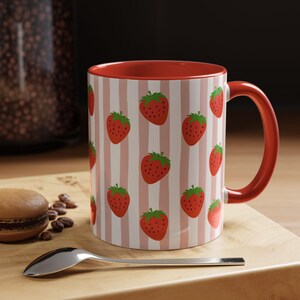 May include: A ceramic mug featuring a repeating pattern of red strawberries on a background of vertical light pink and white stripes. The mug has a solid red interior and a matching red handle, providing a vibrant contrast to the patterned exterior.