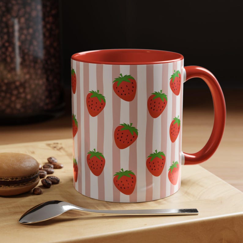 May include: A ceramic mug featuring a repeating pattern of red strawberries on a background of vertical light pink and white stripes. The mug has a solid red interior and a matching red handle, providing a vibrant contrast to the patterned exterior.