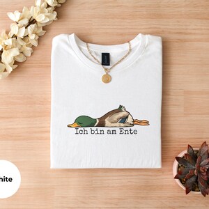 Pode incluir: Uma t-shirt branca com um gr&aacute;fico de um pato-real deitado de barriga para baixo com os olhos fechados. Abaixo do pato, o texto preto diz Ich bin am Ente. Um colar de corrente dourada com um pendente redondo repousa no decote.