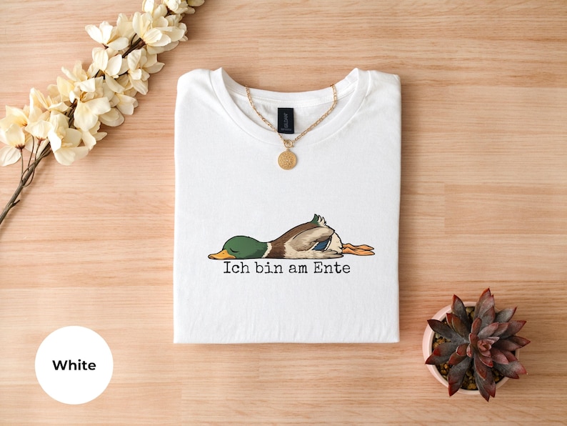 Pode incluir: Uma t-shirt branca com um gr&aacute;fico de um pato-real deitado de barriga para baixo com os olhos fechados. Abaixo do pato, o texto preto diz Ich bin am Ente. Um colar de corrente dourada com um pendente redondo repousa no decote.