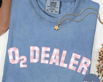Pode incluir: Uma t-shirt azul jeans com gola redonda e etiqueta da marca Comfort Colors. A frente apresenta o texto O2 DEALER num padrão xadrez rosa e branco. Um colar dourado com um pequeno pendente em forma de estrela repousa sobre a gola.