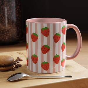 May include: A ceramic mug with a light pink interior and handle, featuring a repeating pattern of red strawberries on a background of vertical white and light pink stripes. The mug has a cylindrical shape and a smooth, glossy finish throughout.