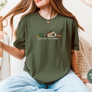 Pode incluir: Uma t-shirt de manga curta verde militar com um gr&aacute;fico de um pato-real deitado de barriga para baixo e o texto "Ich bin am Ente" impresso em letras brancas por baixo da ilustra&ccedil;&atilde;o do pato.