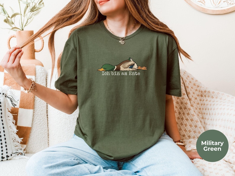 Pode incluir: Uma t-shirt de manga curta verde militar com um gr&aacute;fico de um pato-real deitado de barriga para baixo e o texto "Ich bin am Ente" impresso em letras brancas por baixo da ilustra&ccedil;&atilde;o do pato.