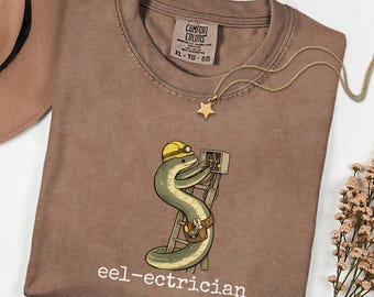 Puede incluir: Una camiseta color espresso con un gráfico de una anguila verde que lleva un casco amarillo, subida a una escalera y trabajando en una caja eléctrica. El texto "eel-ectrician" está impreso en blanco debajo del gráfico.