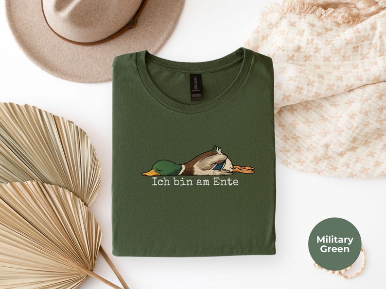 Pode incluir: Uma t-shirt de gola redonda em verde militar com um gr&aacute;fico de um pato-real deitado de barriga para baixo, com o texto Ich bin am Ente impresso em letras brancas por baixo da ilustra&ccedil;&atilde;o do pato na parte da frente da pe&ccedil;a.
