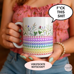 Könnte beinhalten: Ein weißer Keramikbecher mit einer bunten Blumenillustration im oberen Bereich und darunter liegenden Reihen von Kreuzstichmustern in Regenbogenfarben. Der Text F*CK THIS SHIT ist in die unterste Reihe des Musters integriert.