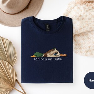 Pode incluir: Uma t-shirt azul-marinho com um gr&aacute;fico de um pato-real deitado de barriga para baixo e o texto Ich bin am Ente impresso em branco por baixo. A pe&ccedil;a tem uma gola redonda e uma etiqueta preta no interior da gola.