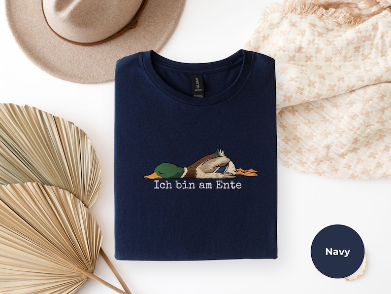 Pode incluir: Uma t-shirt azul-marinho com um gr&aacute;fico de um pato-real deitado de barriga para baixo e o texto Ich bin am Ente impresso em branco por baixo. A pe&ccedil;a tem uma gola redonda e uma etiqueta preta no interior da gola.