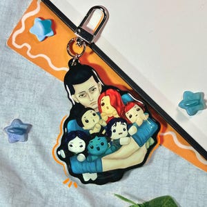 May include: A keychain featuring an illustration of a person holding several small figures. The figures have different hair colors and skin tones. The keychain has a silver clasp and is attached to a notebook.