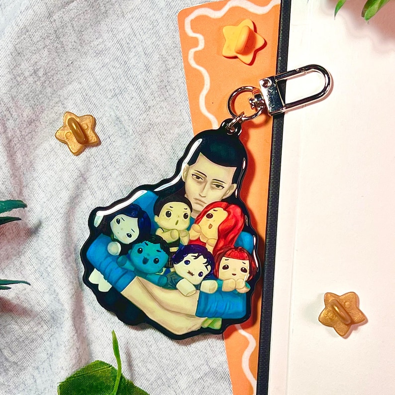 May include: A keychain featuring a cartoon illustration of a person holding several small, colorful figures. The figures have different hair colors and expressions. The keychain has a silver clasp and is set against a light background with star-shaped embellishments.