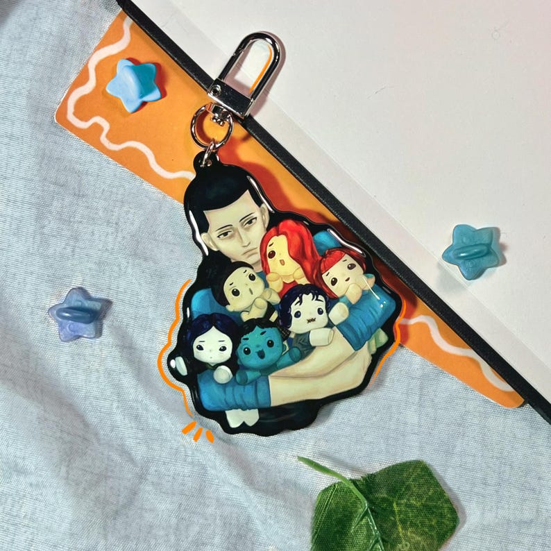 May include: A keychain featuring an illustration of a person holding several small figures. The figures have different hair colors and skin tones. The keychain has a silver clasp and is attached to a notebook.