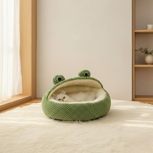May include: A green cat bed shaped like a frog, with two large, round eyes and a textured exterior. The interior is a soft, cream-colored material, and a white cat is nestled inside. The bed is on a white rug.