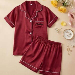 May include: Burgundy satin pajama set with white piping. The short-sleeved top has a notched collar, a breast pocket with the name "Justina" embroidered in black, and button closures. The matching shorts complete the set.