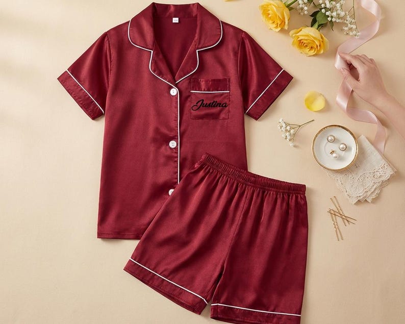May include: Burgundy satin pajama set with white piping. The short-sleeved top has a notched collar, a breast pocket with the name "Justina" embroidered in black, and button closures. The matching shorts complete the set.