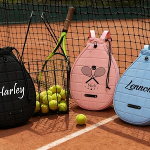 May include: Three tennis-themed backpacks in black, pink, and blue, each with a name on the front. A black backpack has the name "Harley", a pink backpack has "Nick", and a blue backpack has "Lennon". A tennis racket and balls are in the background.