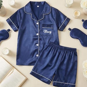 May include: Navy blue satin pajama set with white trim, including a button-down shirt, shorts, and matching sleep masks. The shirt pocket is embroidered with the name "Macy". The set is surrounded by candles, flowers, and an open book.