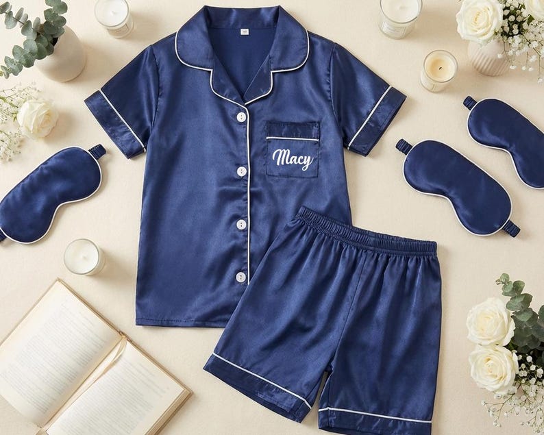 May include: Navy blue satin pajama set with white trim, including a button-down shirt, shorts, and matching sleep masks. The shirt pocket is embroidered with the name "Macy". The set is surrounded by candles, flowers, and an open book.