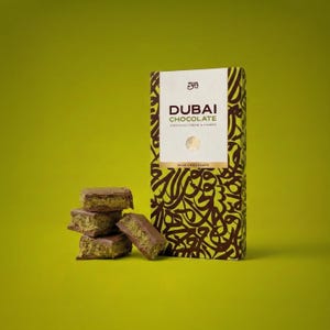 Könnte beinhalten: Eine Schachtel Dubai-Schokolade mit Pistaziencreme und Kunafa sowie mehrere Schokoladenstücke auf grünem Hintergrund. Die Schachtel hat ein weißes Feld mit dem Schriftzug "Dubai Chocolate" in dunkelbrauner Schrift.