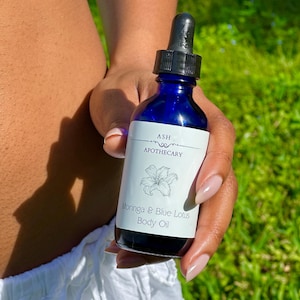May include: A dark blue glass bottle of Moringa & Blue Lotus Body Oil. The bottle has a black dropper and a white label with the text "ASH Apothecary" and a floral illustration. The bottle is held against a background of green grass.