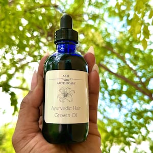 May include: A dark blue glass bottle of Ayurvedic Hair Growth Oil held in a hand. The bottle has a black dropper and a white label with the text "ASH APOTHECARY" and "Ayurvedic Hair Growth Oil". A white flower illustration is also on the label.