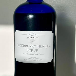 Könnte beinhalten: Eine dunkelblaue Glasflasche mit Holunder-Kräutersirup. Auf dem weißen Etikett steht "ASH APOTHECARY ELDERBERRY HERBAL SYRUP". Gebrauchsanweisungen sind ebenfalls auf dem Etikett aufgedruckt. Die Flasche ist etwa 20 cm hoch.