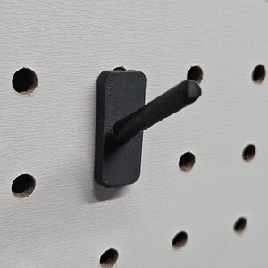 May include: A close-up of a white pegboard with evenly spaced holes. A black pegboard hook is inserted into one of the holes. The hook has a rectangular base and a short, angled arm, designed for hanging items.