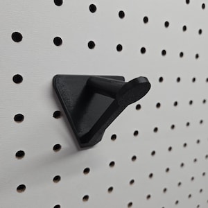 May include: A black pegboard hook with a triangular base and a curved hook. The hook is mounted on a white pegboard with evenly spaced holes. The hook is designed to hold items on the pegboard.