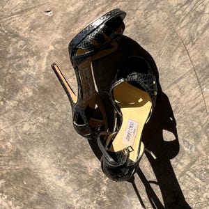 Könnte beinhalten: Ein Paar schwarze High-Heel-Sandalen mit Schlangenmuster. Die Sandalen haben dünne Knöchelriemen und einen Stiletto-Absatz. Der Markenname "JIMMY CHOO" ist auf der Innensohle sichtbar. Die Schuhe stehen vor einem neutralen Hintergrund.