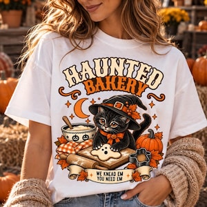 May include: A white t-shirt featuring a graphic of a black cat wearing a witch hat, baking with a rolling pin and a ghost-shaped cookie. The text reads "HAUNTED BAKERY" and "WE KNEAD EM YOU NEED EM" with orange autumn leaves and pumpkins.
