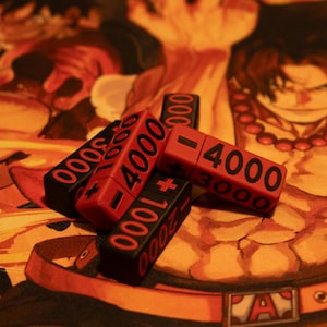 Könnte beinhalten: Eine Ansammlung roter und schwarzer Spielzähler mit Zahlenwerten wie "4000" und "3000" liegt auf einer farbenfrohen, illustrierten Spielmatte. Die Matte zeigt eine Figur mit einer roten Perlenkette und einem Gürtel mit dem Buchstaben "A".