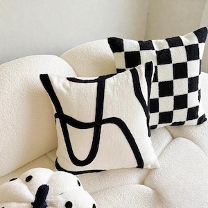 Puede incluir: Dos cojines decorativos con fondo blanco. Uno tiene un diseño abstracto negro y el otro un patrón de tablero de ajedrez en blanco y negro. Los cojines están sobre un sofá blanco texturizado.