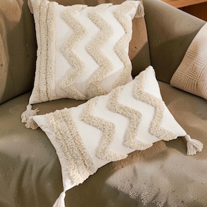Könnte beinhalten: Zwei cremefarbene Zierkissen mit einem strukturierten Zickzackmuster und Fransenquasten. Die Kissen sind rechteckig und quadratisch und liegen auf einem grünen Sofa. Sie verleihen jedem Wohnraum Textur und Stil.