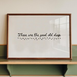 Puede incluir: Una impresión enmarcada con el texto "These are the good old days" en una fuente decorativa, encima de una fila de ilustraciones de estrellas. La impresión se muestra en una estantería verde y beige, montada en una pared blanca. El marco es marrón.