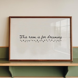 Può includere: Una stampa incorniciata con la scritta "This room is for dreaming" è appoggiata su una mensola verde e beige. La stampa ha una cornice marrone e uno sfondo chiaro. La mensola è montata su un muro bianco.