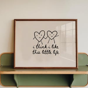 Puede incluir: Un cuadro enmarcado con dos corazones sonrientes tomados de la mano, con el texto "i think i like this little life". El cuadro se exhibe en una estantería verde y de madera.