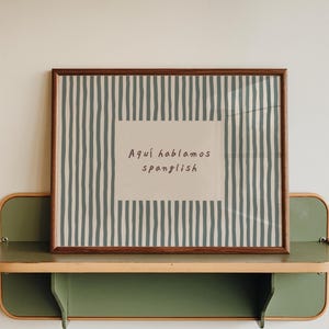 May include: A rectangular framed print featuring a pattern of vertical dark teal stripes on a light background, with a central white rectangle containing the dark text "Aquí hablamos spanglish" in a simple, handwritten-style font.