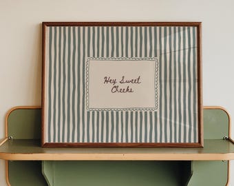 May include: A rectangular framed print featuring a pattern of vertical sage green stripes on a light background. In the center, a white rectangular label with a scalloped border contains the handwritten dark brown text Hey Sweet Cheeks.