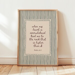 Puede incluir: Una lámina decorativa rectangular en un marco de madera con un panel central beige claro y un borde festoneado. El texto dice: when my heart is overwhelmed lead me to the rock that is higher than I Psalm 61:2. El fondo tiene rayas verticales.