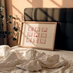 K&ouml;nnte beinhalten: Ein gerahmter Druck mit neun roten Illustrationen von Menschen in Fenstern. Das Kunstwerk hat einen cremefarbenen Hintergrund und einen hellen Holzrahmen. Der Druck wird auf einem Bett mit wei&szlig;er Bettw&auml;sche pr&auml;sentiert.