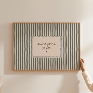 Puede incluir: Un estampado enmarcado con un marco de madera clara. El fondo tiene rayas verticales en una combinación de colores verde apagado y crema. El centro presenta un rectángulo color crema con las palabras "you're gonna go far" y un pequeño corazón.