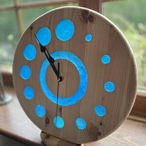 May include: A circular wooden clock with a light brown finish. The clock face has blue circular inlays and a blue inner ring. Black clock hands show the time. The clock is supported by a wooden base.