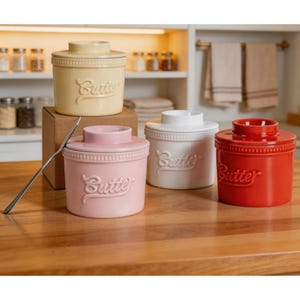 May include: A selection of ceramic butter dishes in shades of yellow, pink, white, and red. Each dish features a lid and the word "Butter" embossed on the front. A silver butter knife is placed on the wooden surface.