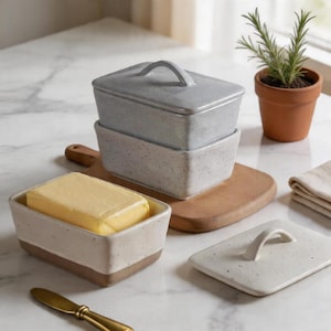 Może przedstawiać: Prostokątne maselniczka z jasnoszarą pokrywką i uchwytem, ​​zawierająca żółty blok masła. Naczynie stoi na drewnianej desce. Na zdjęciu widać również małą roślinę doniczkową i nóż.