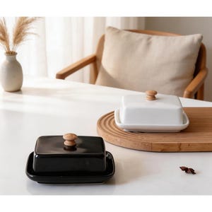 Puede incluir: Dos mantequilleras de cerámica, una negra y otra blanca, cada una con un pomo de madera en la tapa. El plato negro se asienta sobre una base a juego, mientras que el blanco descansa sobre una tabla de servir de madera. Un jarrón con hierba seca en el fondo.