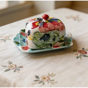 Puede incluir: Una mantequera cuadrada de cerámica con diseño floral. La tapa tiene un pomo rojo y está decorada con flores rojas, rosas, azules y verdes. La base es una placa rectangular azul claro. El plato está sobre un mantel crema.