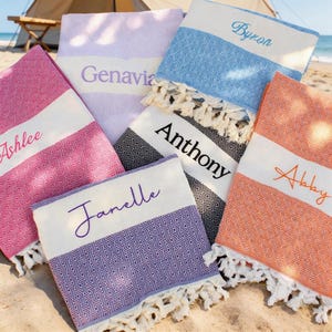 May include: A selection of patterned beach towels in various colours, including pink, blue, and orange, with white fringe. Each towel features a white band with a name embroidered: Ashlee, Genavia, Byron, Anthony, Abby, and Janelle. The towels are on a sandy beach.
